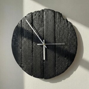 May include: A round, black wooden wall clock with white hands. The clock face is made of vertical wooden planks, giving it a rustic appearance. The hands are thin and white, contrasting against the dark background. The clock is mounted on a white wall.