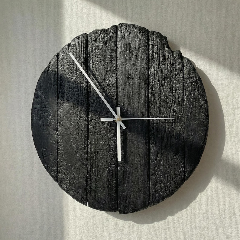 May include: A round, black wooden wall clock with white hands. The clock face is made of vertical wooden planks, giving it a rustic appearance. The hands are thin and white, contrasting against the dark background. The clock is mounted on a white wall.