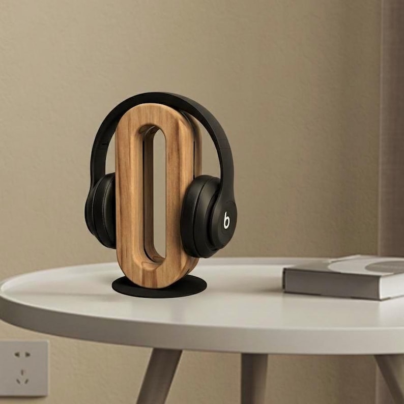 May include: A wooden headphone stand in a unique O-shape, holding a pair of black headphones. The stand is made of light-colored wood and sits on a black base. The headphones have a curved design.