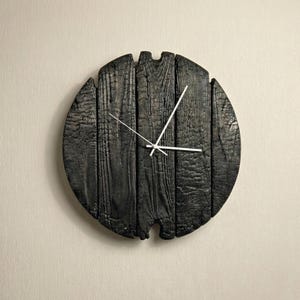 May include: A round, black wooden clock with white hands. The clock face is made of charred wood planks, giving it a rustic appearance. The clock is mounted on a light beige wall.