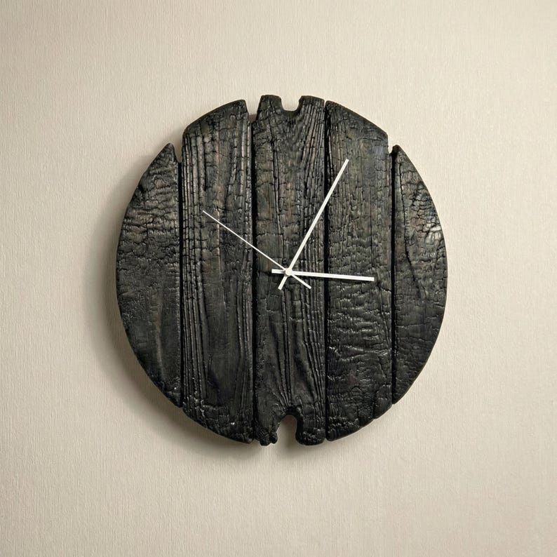 May include: A round, black wooden clock with white hands. The clock face is made of charred wood planks, giving it a rustic appearance. The clock is mounted on a light beige wall.