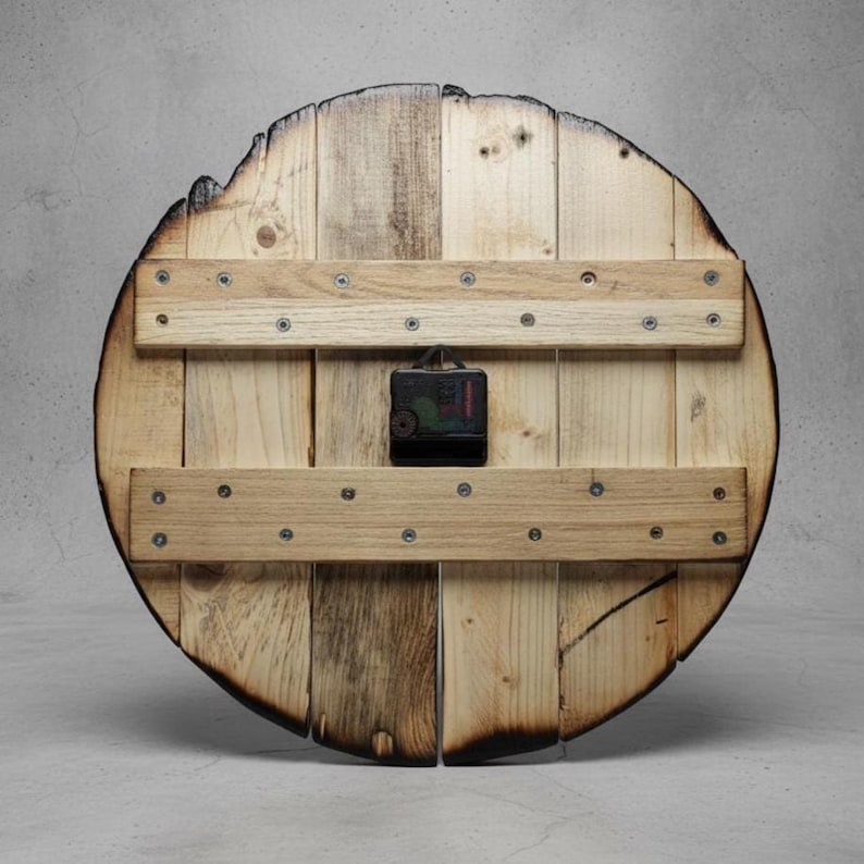May include: A round wooden clock with a rustic design. The clock face is made of light-colored wooden planks, with darker, charred edges. Two horizontal wooden supports are attached to the back with screws, and a black clock mechanism is centered.