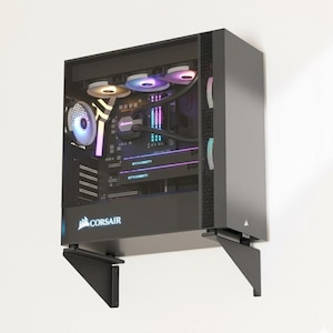 May include: A black computer case mounted on a white wall. The case has a transparent side panel revealing internal components, including fans with colorful LED lighting. The Corsair logo is visible on the front. The case is supported by two black brackets.