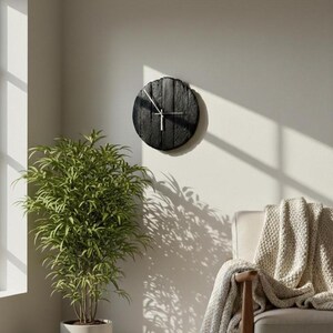 May include: A round, black wooden wall clock with silver hands hangs on a light-colored wall. The clock has a rustic, textured appearance. The image also shows a green plant in a white pot and a chair with a beige blanket.