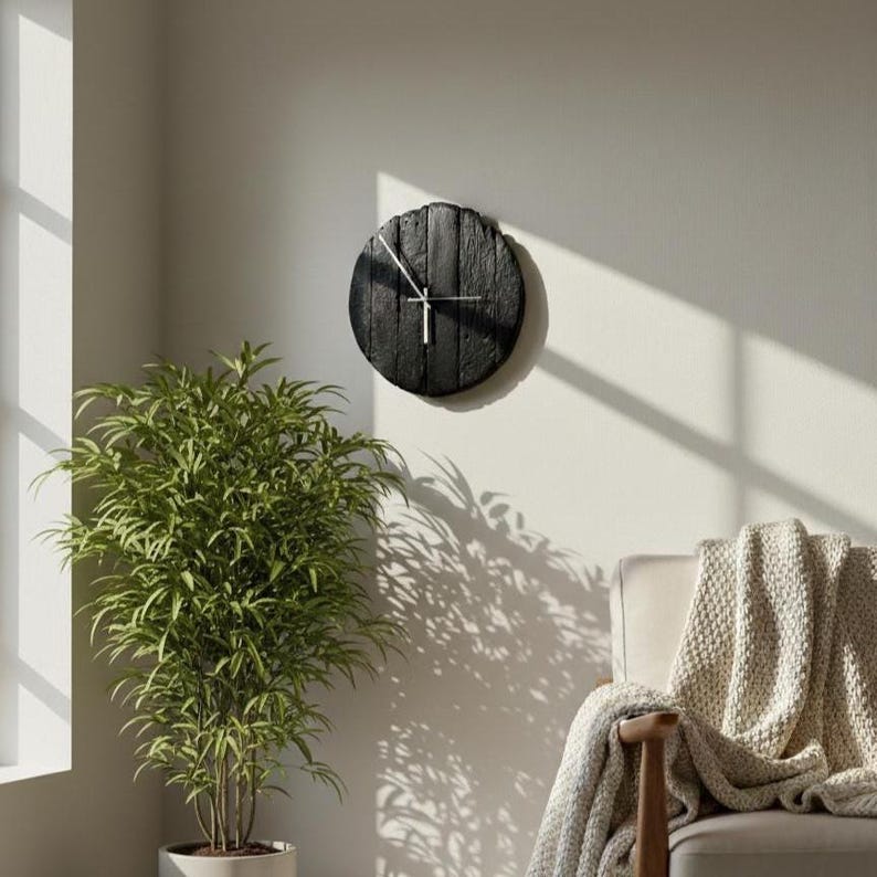 May include: A round, black wooden wall clock with silver hands hangs on a light-colored wall. The clock has a rustic, textured appearance. The image also shows a green plant in a white pot and a chair with a beige blanket.
