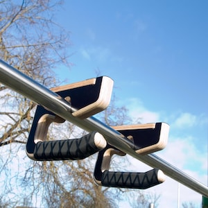 以下が含まれることがあります： 金属製のバーに取り付けられた、黒と木製のプッシュアップバーのペア。 プッシュアップバーは黒いグリップと木製のベースを備えています。 背景には青い空と木が表示されています。