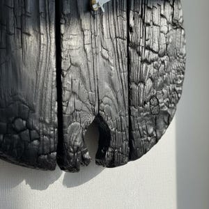 May include: Close-up of a round, black wooden clock. The clock face is made of three vertical wooden planks with a charred, textured surface. The clock hands are white and the clock is mounted on a white wall.