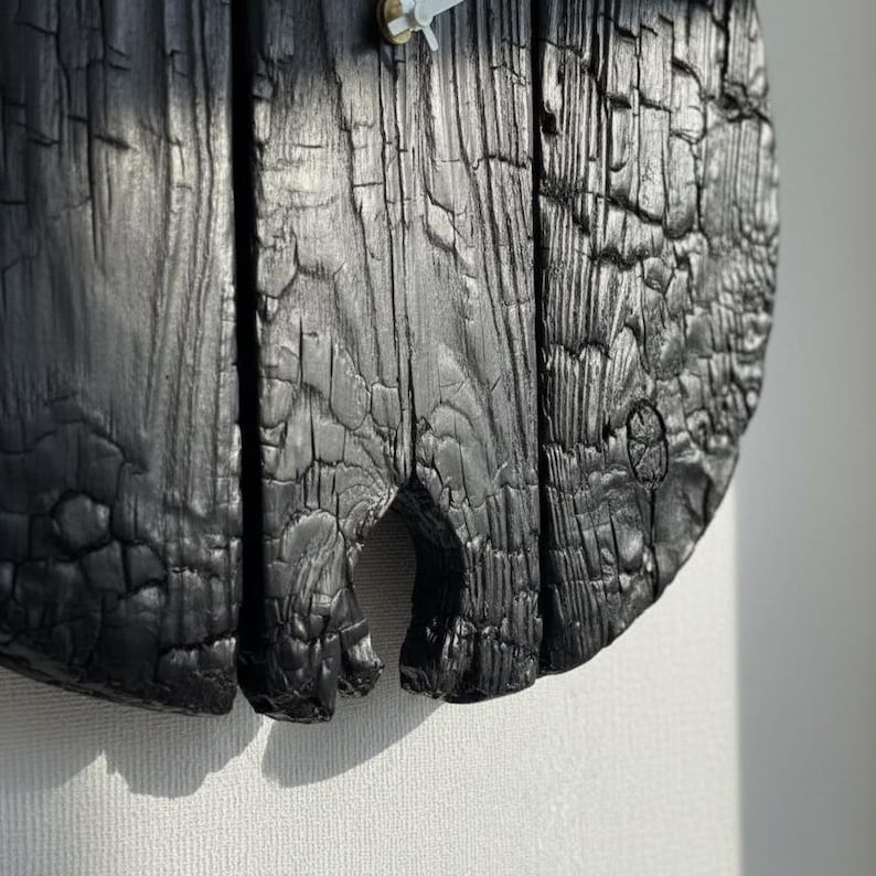 May include: Close-up of a round, black wooden clock. The clock face is made of three vertical wooden planks with a charred, textured surface. The clock hands are white and the clock is mounted on a white wall.