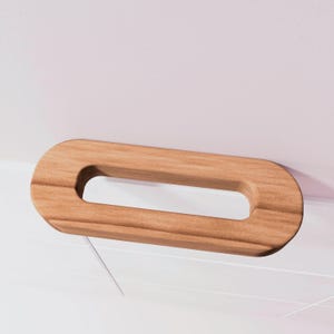 May include: A wooden cabinet pull with an oval shape and a rectangular opening in the center. The pull is made of light brown wood and is mounted on a white surface.
