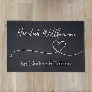 May include: A rectangular black doormat with white script that reads "Herzlich Willkommen" above a white heart and a curved line. Below the heart, the text reads "bei Nadine & Fabian".