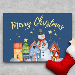 Puede incluir: Una tarjeta azul con el texto "Merry Christmas" en letras doradas. La tarjeta presenta una ilustración de acuarela de un muñeco de nieve de pie frente a una fila de casas con un árbol de Navidad. Las casas están pintadas en tonos de azul, rojo y amarillo.
