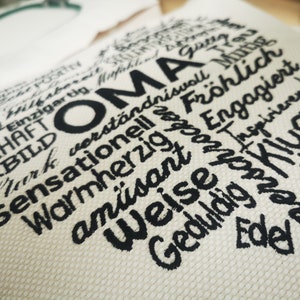 May include: A white fabric heart-shaped pillow with black embroidered text. The text reads "OMA" and includes words like "warmherzig", "am&uuml;sant", "verst&auml;ndnisvoll", "fr&ouml;hlich", "toll", and "weise".
