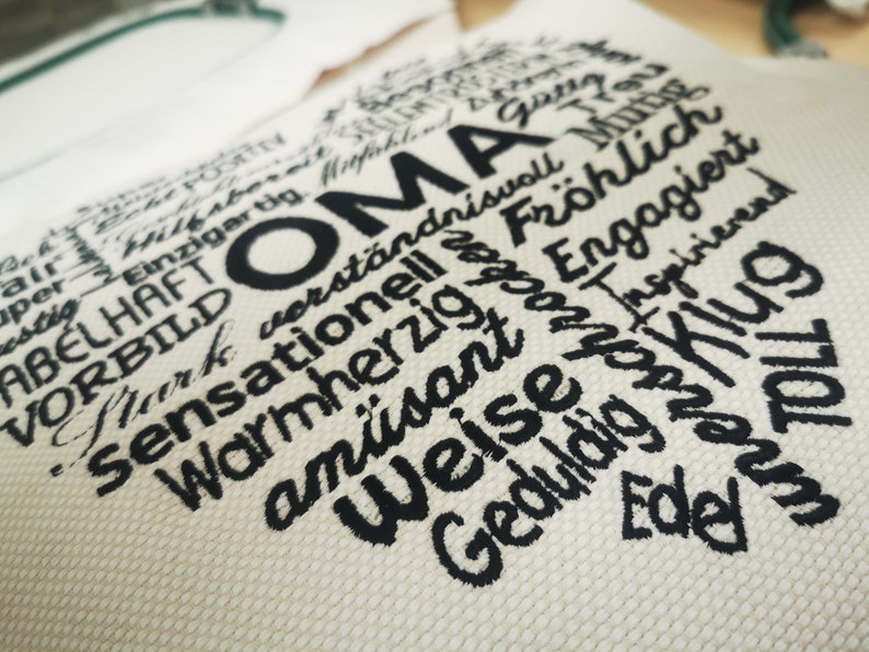 May include: A white fabric heart-shaped pillow with black embroidered text. The text reads "OMA" and includes words like "warmherzig", "am&uuml;sant", "verst&auml;ndnisvoll", "fr&ouml;hlich", "toll", and "weise".