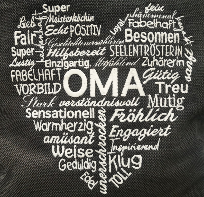 May include: A black heart-shaped canvas bag with white embroidery. The words "OMA" are in the center of the heart, surrounded by positive adjectives like "Super", "Fair", "Hilfsbereit", "Treu", "Fr&ouml;hlich", "Klug", and "TOLL".
