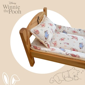 May include: A wooden doll bed featuring a light-coloured frame and bedding decorated with a Winnie the Pooh character print. The set includes a matching pillow and a mattress pad with a soft, cream-coloured fleece lining along the edges.