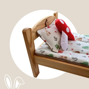 May include: A small wooden bed frame with a light natural finish, featuring a white pillow and mattress cover printed with small bears, trees, and mushrooms. A plush red mushroom-shaped toy with white spots rests on the bed surface.