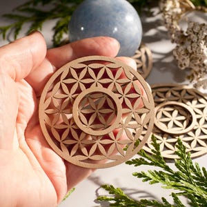 Könnte beinhalten: Ein Untersetzer aus Holz mit einem geometrischen Muster, bekannt als Blume des Lebens, in hellbrauner Farbe. Der Untersetzer wird in einer Hand gehalten.