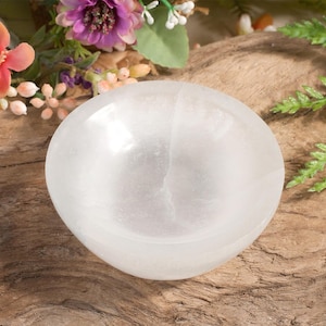 May include: A white selenite bowl, also known as a sound bowl, is shown on a wooden surface. The bowl is smooth and has a slight sheen. The bowl is surrounded by green and purple flowers.