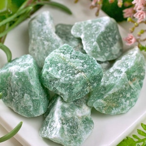 May include: A collection of rough, green aventurine stones on a white plate, accented by greenery and pink flowers. The stones have a textured surface and varying shades of green, with some white veining.