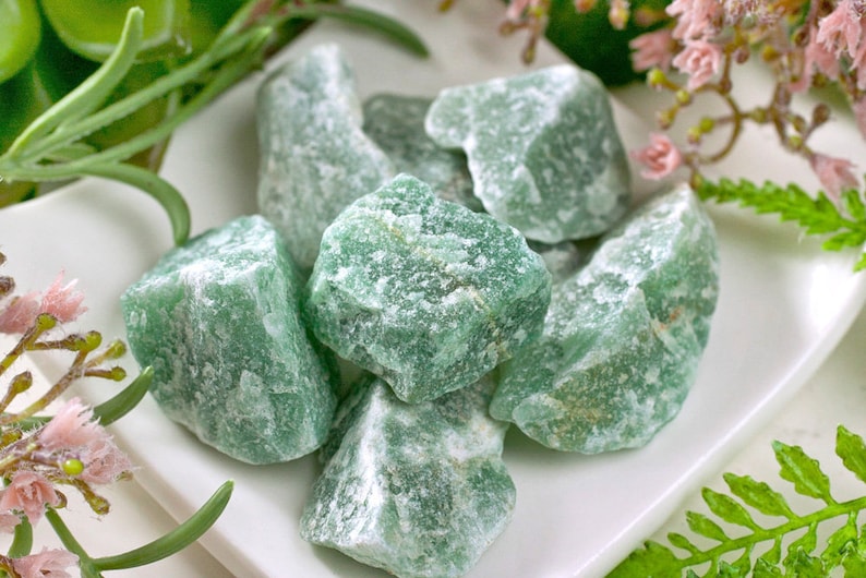 May include: A collection of rough, green aventurine stones on a white plate, accented by greenery and pink flowers. The stones have a textured surface and varying shades of green, with some white veining.
