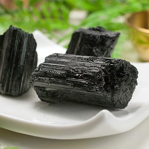 May include: Three black tourmaline crystals on a white ceramic dish. The crystals are raw and have a striated texture.