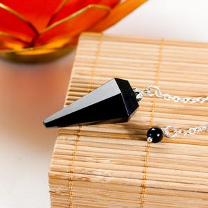 Könnte beinhalten: Ein schwarzes, facettiertes Edelsteinpendel mit spitzem Ende, das an einer silberfarbenen Metallkette befestigt ist. Die Kette weist in der Nähe des Verbindungspunkts eine kleine, runde schwarze Perle auf, die einen Kontrast zur polierten Oberfläche bildet.