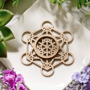 Könnte beinhalten: Ein hölzerner Metatron-Würfel, ein geometrisches Symbol, ist auf einer weißen quadratischen Fliese zentriert. Das komplizierte Design weist miteinander verbundene Kreise und Linien auf. Das Bild ist von grünen Sukkulenten, lila Blumen und Grün umgeben.