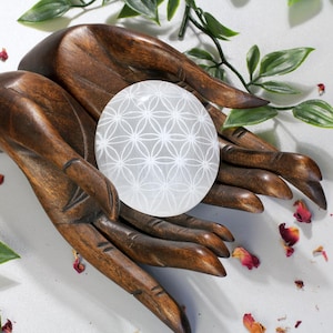 Può includere: Una sfera di cristallo di selenite bianca con un disegno a fiore della vita, che riposa su un paio di mani di legno intagliate.