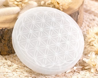 May include: A white selenite charging plate featuring a carved flower of life design. The plate is round and has a smooth, polished surface.