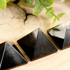 May include: Three black obsidian pyramids of varying sizes on a light wood surface.