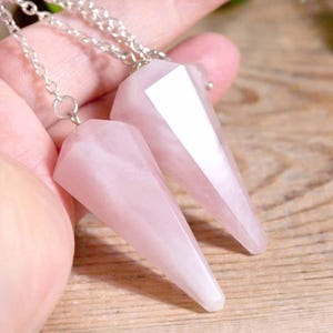 May include: Two pink rose quartz crystal pendulums with silver chains. The pendulums are faceted and cone-shaped, with a polished finish. The background is a wooden surface.