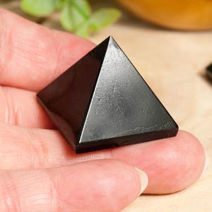 May include: A black obsidian pyramid crystal, a popular choice for grounding and protection, is held in a hand. The pyramid is smooth and polished, with a sharp point at the top.