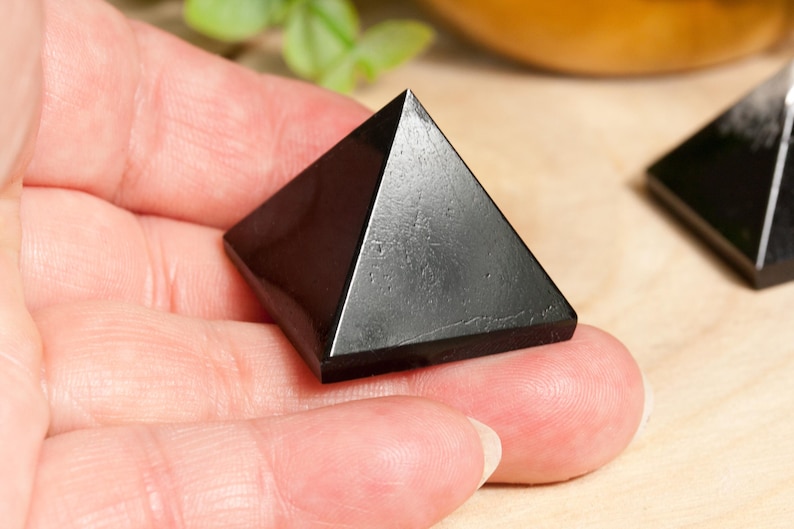 May include: A black obsidian pyramid crystal, a popular choice for grounding and protection, is held in a hand. The pyramid is smooth and polished, with a sharp point at the top.