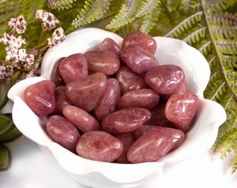 Può includere: Una ciotola bianca smerlata piena di pietre preziose lucide, rosa-rosse. Le pietre hanno una forma liscia e arrotondata e variano di dimensioni. Felci verdi e accenti floreali bianchi sullo sfondo.