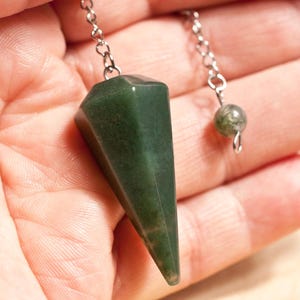Könnte beinhalten: Ein dunkelgrünes, facettiertes Pendel mit einer silbernen Kette und einer kleinen, runden Perle. Das Pendel wird in einer Hand gehalten, wodurch seine polierte Oberfläche und geometrische Form zur Geltung kommen. Kette und Perle bilden einen zarten Kontrast zum Stein.