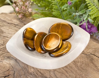 May include: Five smooth, polished tiger's eye stones in a white selenite bowl. The stones are various shades of brown and gold with a shimmering, chatoyant effect.
