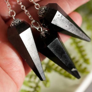 Könnte beinhalten: Drei schwarze Kristallpendel mit silbernen Ketten und dekorativen Kappen. Die Pendel werden in einer Hand gehalten und zeigen ihr facettiertes, spitzes Design. Die Kristalle haben eine polierte, reflektierende Oberfläche.
