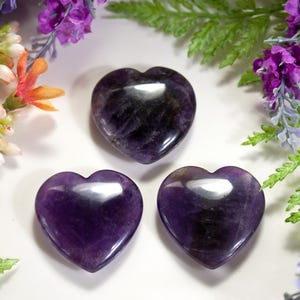 Può includere: Tre cristalli di ametista a forma di cuore di colore viola su uno sfondo bianco. I cristalli sono lucidati e hanno una superficie liscia e lucida.
