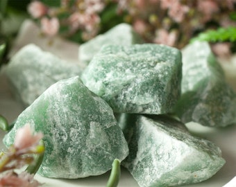 May include: A close-up of several rough-cut, green aventurine stones. The stones have a textured, crystalline appearance and vary in size. The background features soft-focus greenery and pink flowers, creating a natural aesthetic.