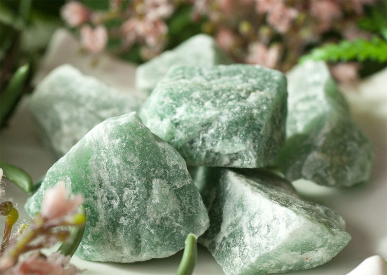 May include: A close-up of several rough-cut, green aventurine stones. The stones have a textured, crystalline appearance and vary in size. The background features soft-focus greenery and pink flowers, creating a natural aesthetic.