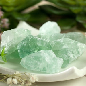 Può includere: Una collezione di cristalli di fluorite verde su un piatto bianco. I cristalli sono grezzi e hanno un aspetto naturale non levigato.