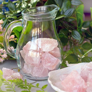 May include: A glass pitcher filled with pink rose quartz crystals. The pitcher is sitting on a white surface with more rose quartz crystals scattered around it.