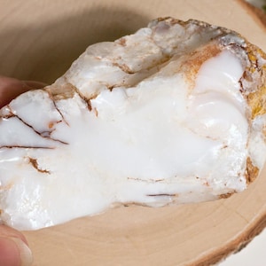 Könnte beinhalten: Ein rohes, unpoliertes weißes Opal-Exemplar mit einer glatten, glänzenden Oberfläche, sichtbaren braunen und lohfarbenen Mineraleinschlüssen und natürlichen Rissen im Stein, das ein milchiges, durchscheinendes Aussehen mit erdigen Tönen aufweist.