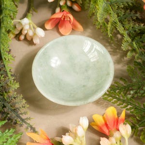 Könnte beinhalten: Eine kleine, hellgrüne Schale mit glatter Oberfläche ist von grünem und braunem Laub sowie orangefarbenen und weißen Blüten umgeben. Die Schale ist in der Mitte des Bildes zentriert, mit den Blumen und dem Laub darum angeordnet.