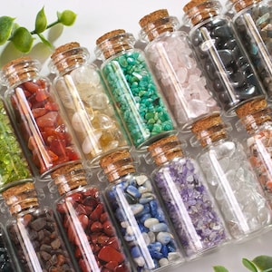 May include: A collection of small glass bottles filled with various types of colourful crystals and stones. The bottles have cork stoppers and are arranged in a row on a white surface.