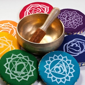 Op de afbeelding: Zeven kleurrijke ronde kussens met verschillende symbolen en een gouden zangkom met een houten slaagstok. De kussens zijn in een cirkel rond de kom gerangschikt.