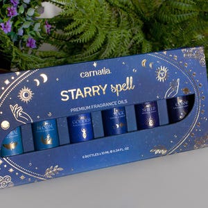 Könnte beinhalten: Ein marineblaues Set mit Premium-Duftölen mit der Aufschrift "Starry Spell" und goldenen Himmelsmotiven. Das Set enthält sechs 10 ml Flaschen mit Namen wie "Bloom" und "Goddess". Die Box ist mit Monden, Sternen und Handillustrationen verziert.