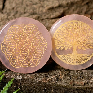 Könnte beinhalten: Zwei runde Untersetzer aus rosa Rosenquarz mit goldenen, geätzten Designs. Ein Untersetzer zeigt ein Lebensblume-Design und der andere ein Lebensbaum-Design.