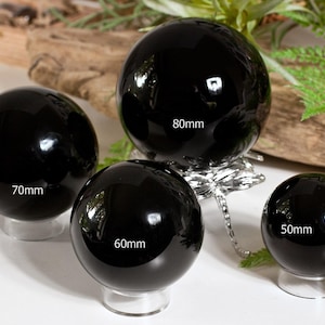 Op de afbeelding: Vier glanzende zwarte bollen van verschillende afmetingen, variërend van 50 mm tot 80 mm in diameter, worden gepresenteerd op transparante standaards. De bollen reflecteren licht. De achtergrond bevat groen en een stuk drijfhout.