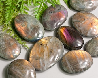Op de afbeelding: Een verzameling van negen ovale labradorit palmstenen. De stenen zijn een mix van grijs, wit en bruin met iriserende flitsen van paars, oranje en goud.
